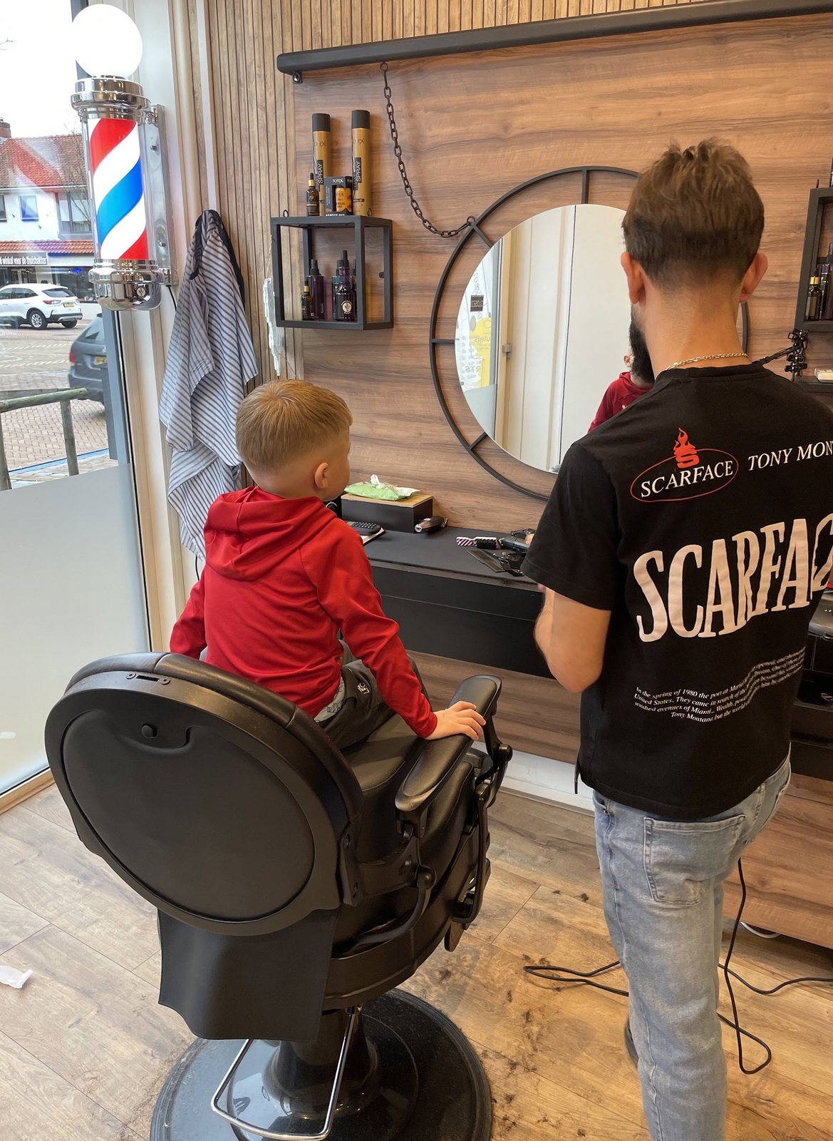 Jongen in de barberstoel, van achteren gezien, met de kapper aan de spiegel