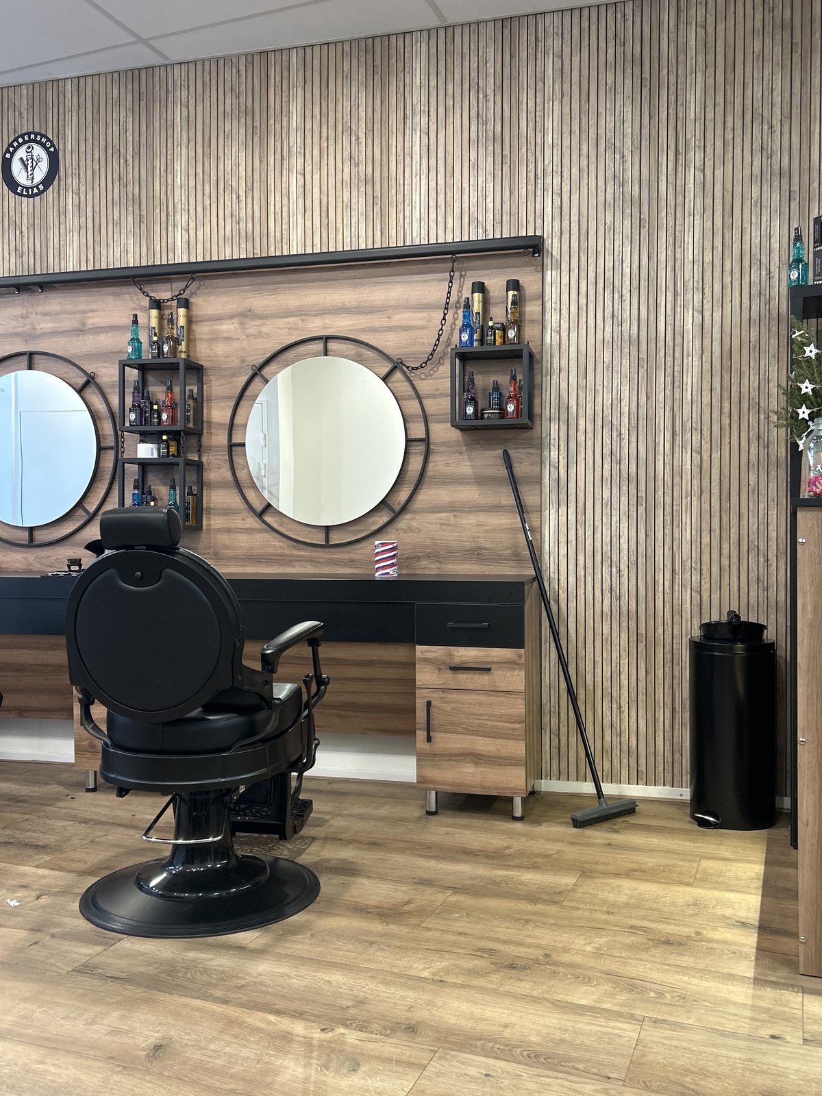 Zwarte barberstoel voor twee ronde spiegels aan een wand van eikenhouten lamellen